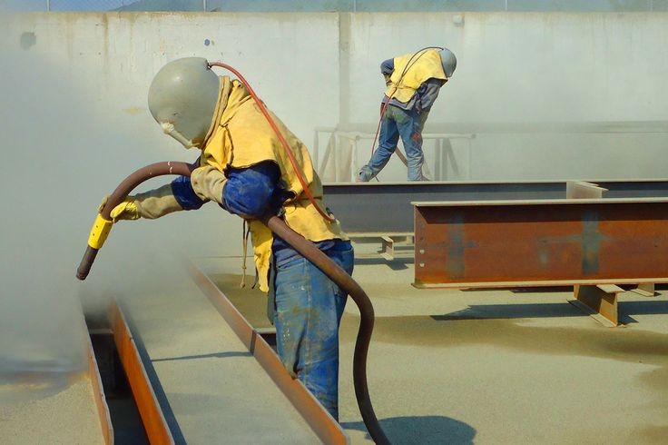 Opérateurs équipés de casques de protection effectuant le sablage de poutres métalliques en acier.